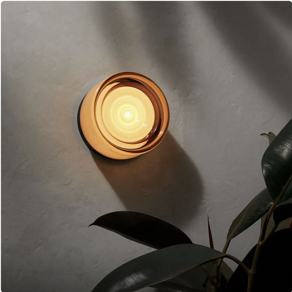 Wenche Studios Ahni wall sconce lit on a plaster wall with plant shadow